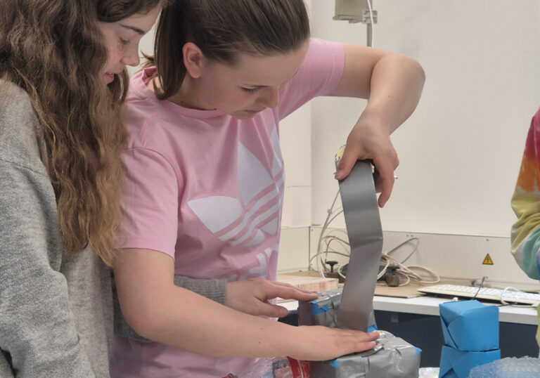 Zwei Mädchen arbeiten an ihrem Projekt. Ein Mädchen klebt etwas mit silbernen Gewebeband.