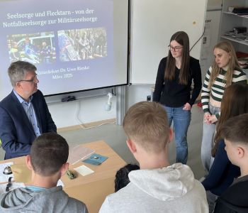 Ein Mann sitzt an einem Tisch. Eine Gruppe Schülerinnen und Schüler umringt ihn.